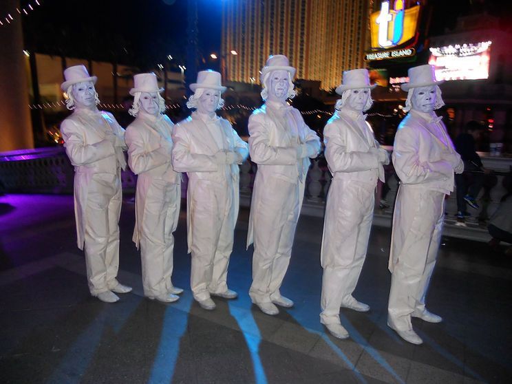 Seven performers in white suits, hats, and masks pose outdoors at night.
