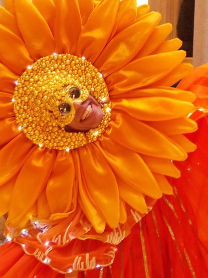 Person in a sunflower costume, smiling. Bright orange petals with sparkly center.