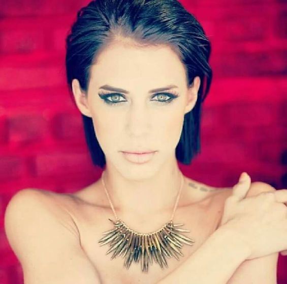 Woman with dark hair, wearing necklace, looking at the viewer against a red background.
