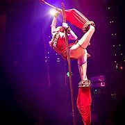 Acrobat performs on a vertical pole, draped in red fabric, lit by spotlights, stage setting.