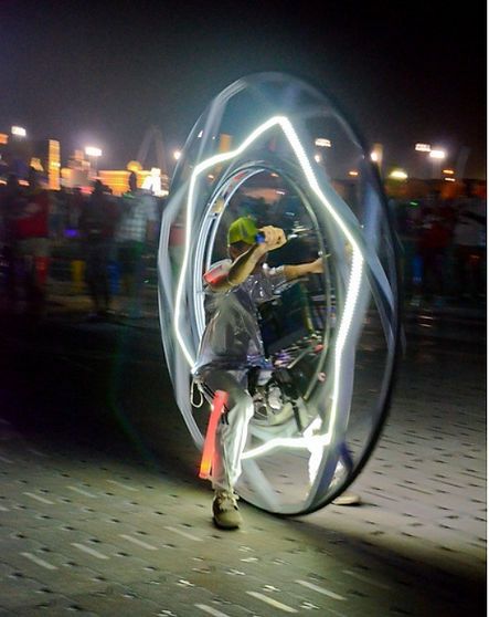 Person riding a glowing, circular vehicle at night in a crowd; streaks of light highlight the rider and structure.