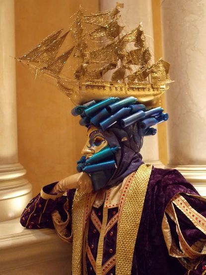 Person in elaborate Venetian mask and costume with a ship-shaped headdress, leaning on a pillar.