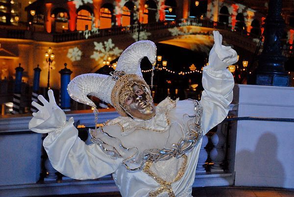 White-costumed performer in a Venetian jester mask, gesturing, against a backdrop of festive lights and architecture.