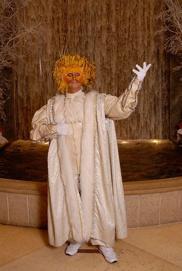 Person in cream-colored costume and mask gestures near a water fountain.