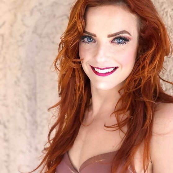 Woman with red hair and blue eyes smiles, wearing makeup and a mauve top, posing in front of a textured wall.
