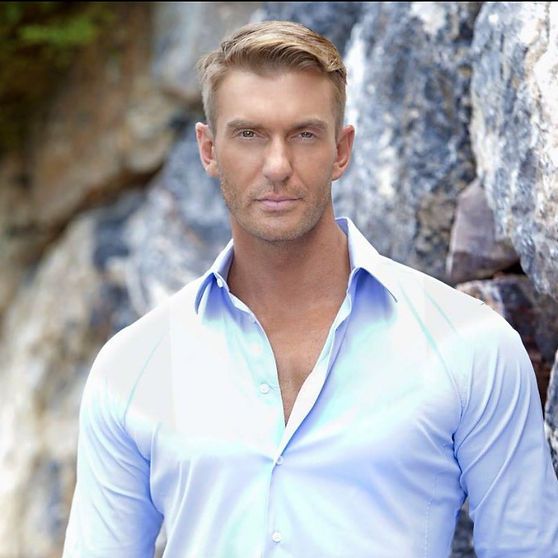 Blond man in a light blue button-down shirt against a rocky backdrop, looking directly at the viewer.