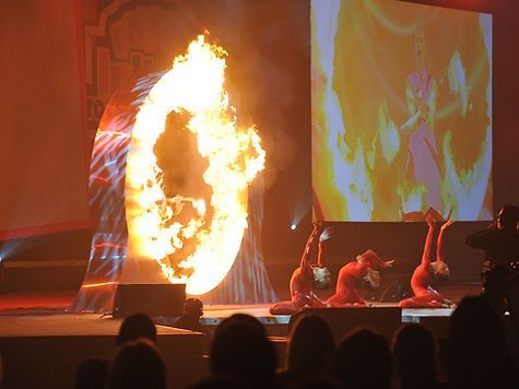 Three acrobats in red suits perform near a fiery ring on stage, with a projection in the background.