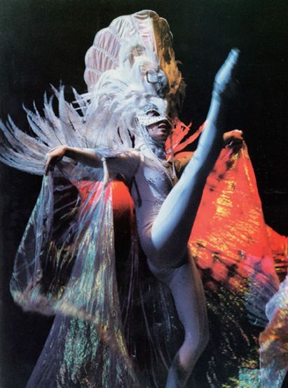 Ballet dancer in white costume, legs extended, performing on stage with elaborate headdress and cape.