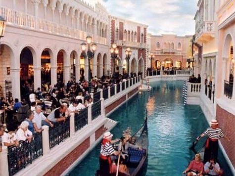 A canal scene with gondolas, restaurants, and people in a Venetian-style setting.