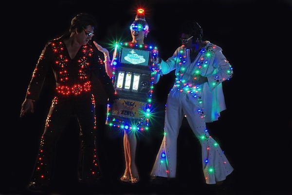 Three people in Elvis costumes covered in colorful lights; one holds a slot machine.