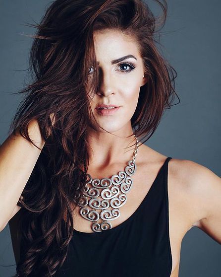 Woman with brown hair wearing a black dress and silver necklace, looking at the camera.