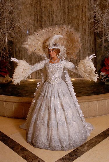 Woman in elaborate white dress with feathered accents, arms outstretched, in front of a fountain.