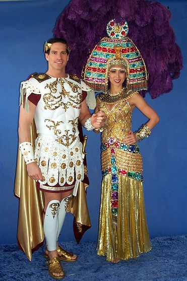 Man in Roman armor and woman in gold Egyptian attire pose. Blue background.