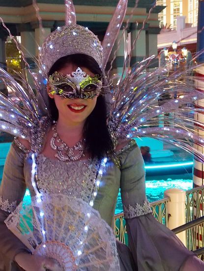 Woman in silver Venetian carnival costume with mask and light-up wings, posing near water.