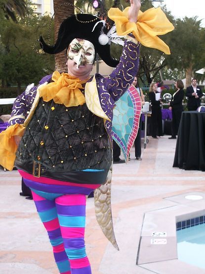 Person in a colorful jester costume with a mask, outdoors by a pool, waving.