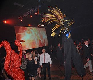 A group of people are dancing in a room with a statue of a swan on stilts.