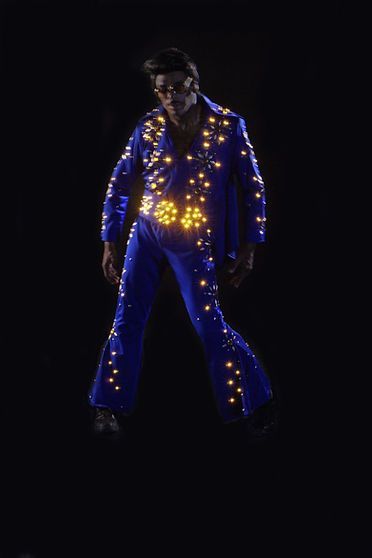 Man in a blue jumpsuit with glowing embellishments, posed like Elvis on a dark stage.