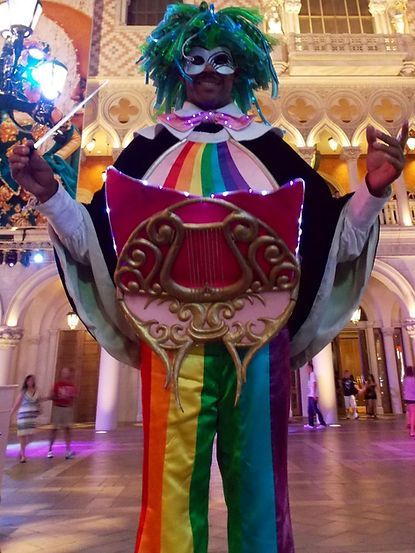 Person in a colorful rainbow outfit with a mask and harp costume posing in a grand interior.