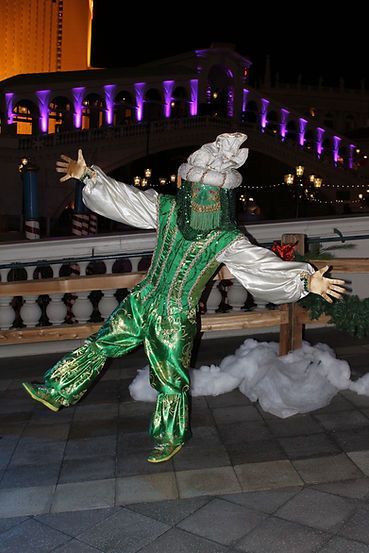 Person in a green costume with a box head, performing outdoors with a bridge in the background.