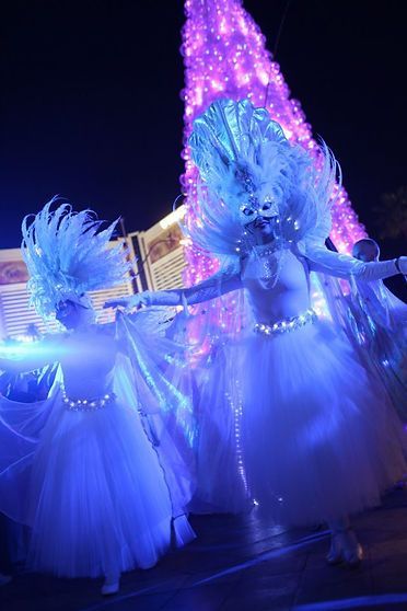 Two dancers in white gowns and headdresses perform in front of a purple lit tree.