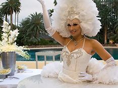 Woman in white feathery costume raises arm, posing by a pool, outdoor setting.