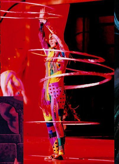 A performer in colorful costume spins multiple hoops around their body on a stage bathed in red light.