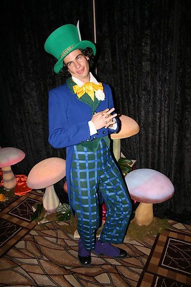 Man in Mad Hatter costume, wearing green top hat, blue coat, plaid pants; standing with mushroom props.