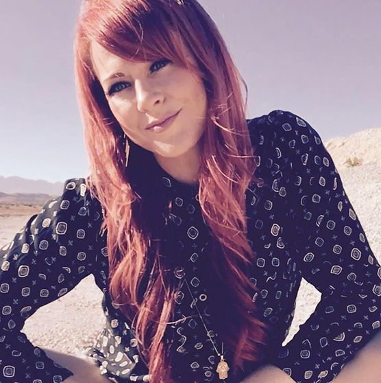 Woman with red hair smiling outdoors, wearing a black patterned shirt.