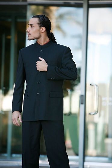 Man in black suit, ponytail, looking to the side, standing in front of glass doors.