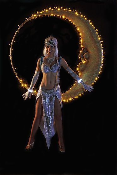 Woman in silver costume, posing in front of a crescent moon lit with fairy lights.