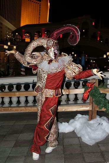 A person in a jester costume with arms outstretched, outdoors near a Christmas decoration.