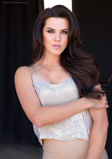 Woman with dark hair in a silver top and beige skirt, posing with one arm across chest, against a black background.