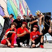 Group of eight people posing in front of a colorful mural. They wear casual clothing and strike confident poses.