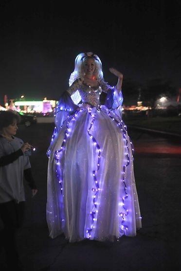 Woman in illuminated gown on stilts at night, waving.