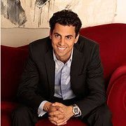 Man in a black suit, light blue shirt, and silver watch, smiling on a red couch.