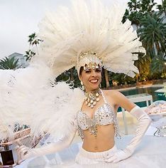 Woman in elaborate white feather headdress and costume, smiling near a pool.