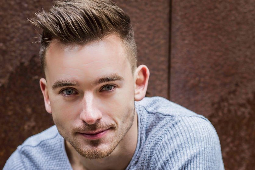 Man with short brown hair and light blue eyes smiles at the camera against a rusted metal backdrop.