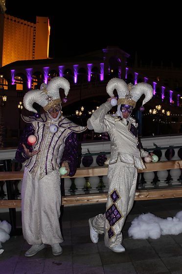 Two performers in white, horned jester costumes juggle colorful balls outdoors at night.