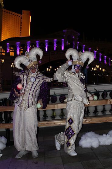 Two performers in white and purple jester costumes juggle glowing balls outdoors at night.
