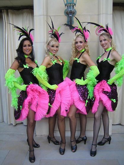 Four women in stage costumes: black tops, pink ruffled skirts, green sleeves, and feathered headpieces, posing outdoors.