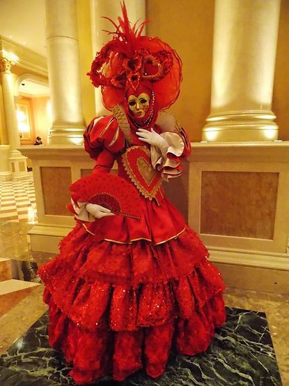 Person in elaborate red costume with mask, feather headdress, and tiered skirt stands by pillars.