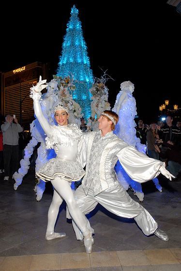 Dancers in silver and white costumes pose in front of a lit blue Christmas tree at night.