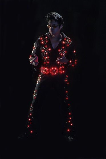 A man in a dark jumpsuit with red glowing accents, possibly an Elvis impersonator, performs on stage.