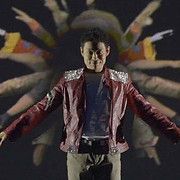 Man in red jacket with outstretched arms, surrounded by blurred dancers in a stage setting.