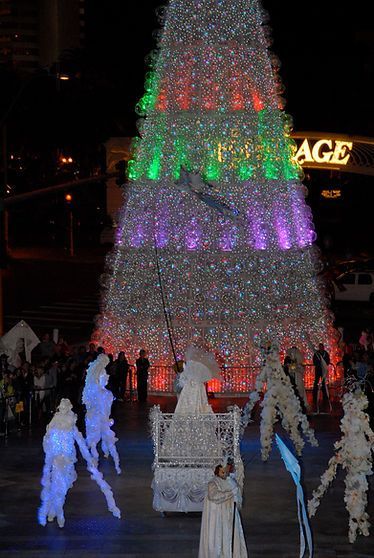 Holiday performers in white costumes with blue accents dance in front of a tall, illuminated Christmas tree at night.