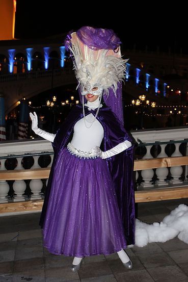 Person in purple costume poses outdoors, near a bridge. They wear a large feathered headdress, cape, and gloves.
