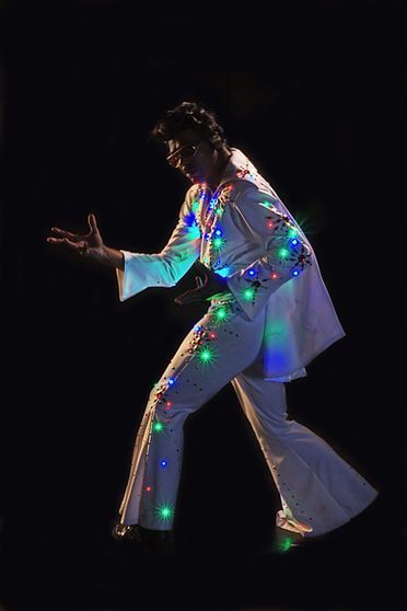 Elvis impersonator in white suit with embedded colorful lights, striking a pose against a black background.