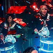 Two drummers performing on illuminated drums, surrounded by red confetti.