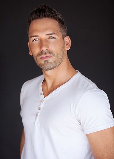 Man in a white V-neck shirt poses with a serious expression against a dark background.