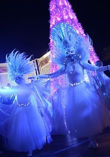 A group of women in white dresses are dancing in front of a christmas tree.